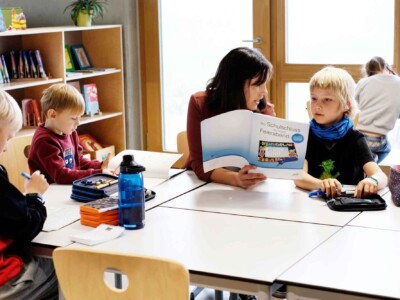 Eine Lehrerin sitzt mit drei Schülern am Tisch und bespricht mit ihnen den Inhalt eines Heftes.