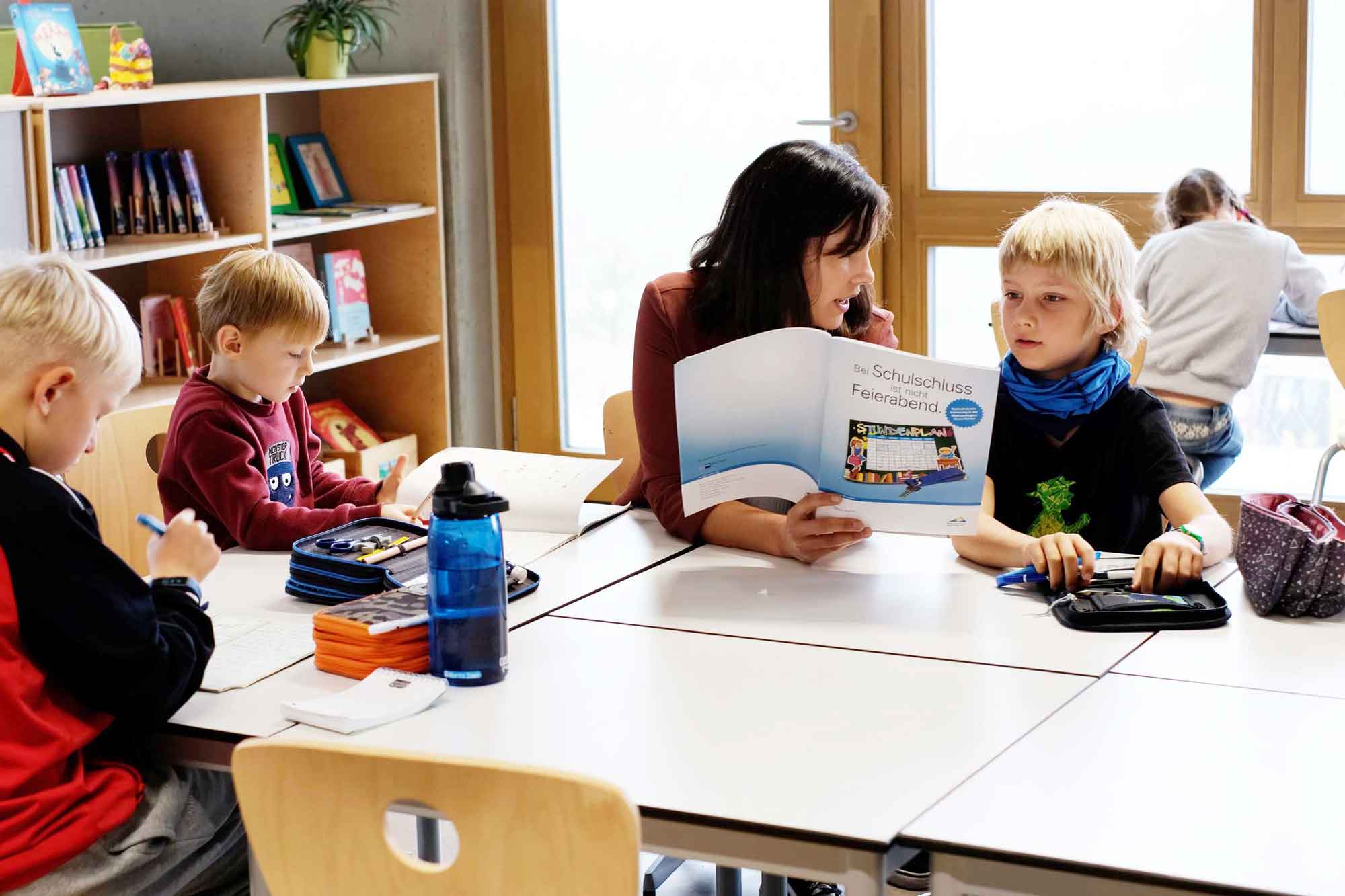 Eine Lehrerin sitzt mit drei Schülern am Tisch und bespricht mit ihnen den Inhalt eines Heftes.