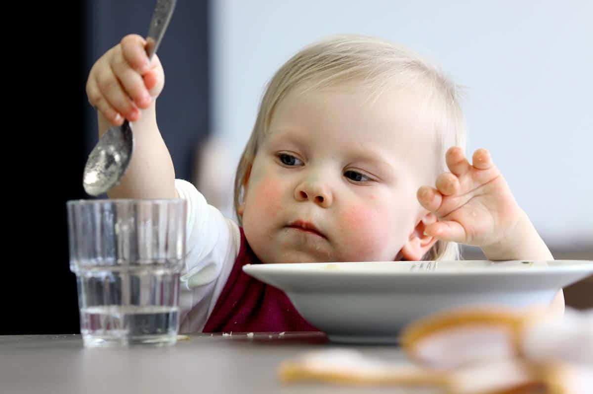 Blondes Kind hebt einen Löffel über ein Glas. Vor dem Kind steht ein Suppenteller.