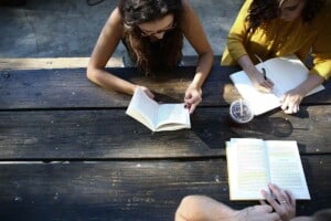Drei Personen sitzen an einer Picknickbank. Zwei Personen lesen ein Buch, eine Frau schreibt in ein Heft.