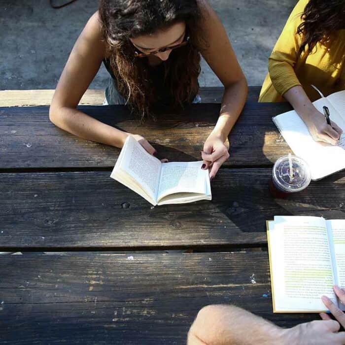 Drei Personen sitzen an einer Picknickbank. Zwei Personen lesen ein Buch, eine Frau schreibt in ein Heft.