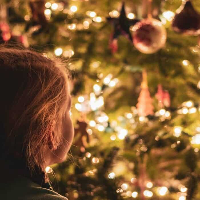 Ein Kind steht vor einem geschmückten Weihnachtsbaum mit Kugeln und Lichterketten.