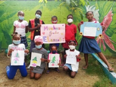 Neun Kinder stehen vor einer grüner Wand. Die Kinder halten Blätter mit einem Tannenbaum. Dahinter halten Kinder ein rosa Plakat, auf dem steht: Muchas Gracias Element-i.