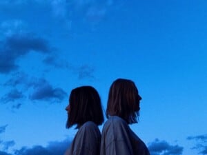 Zwei junge Frauen stehen Rücken an Rücken. Über ihnen sieht man einen blauen Himmel mit Wolken.