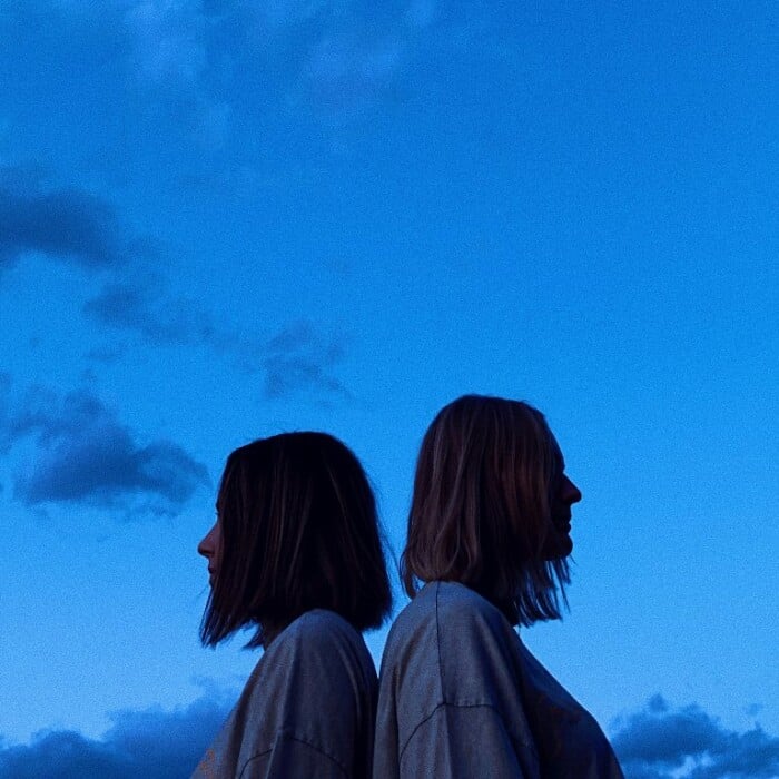 Zwei junge Frauen stehen Rücken an Rücken. Über ihnen sieht man einen blauen Himmel mit Wolken.