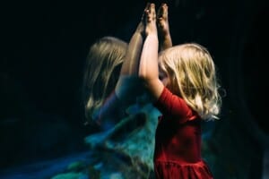 Ein Mädchen mit blonden Haaren und im roten Kleid hält die Hände nach oben an ein Aquariumsglas. Das Mädchen spiegelt sich im Glas.