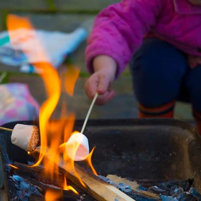 Ein Kind mit einer pinken Jacke, blauen Hose und Gummistiefeln hält ein Marshmallow an einem Stab in ein Lagerfeuer. Ein zweites Marshmallow wird von links in das Feuer gehalten.