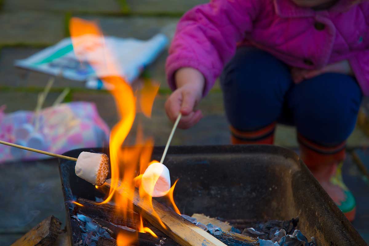 Ein Kind mit einer pinken Jacke, blauen Hose und Gummistiefeln hält ein Marshmallow an einem Stab in ein Lagerfeuer. Ein zweites Marshmallow wird von links in das Feuer gehalten.