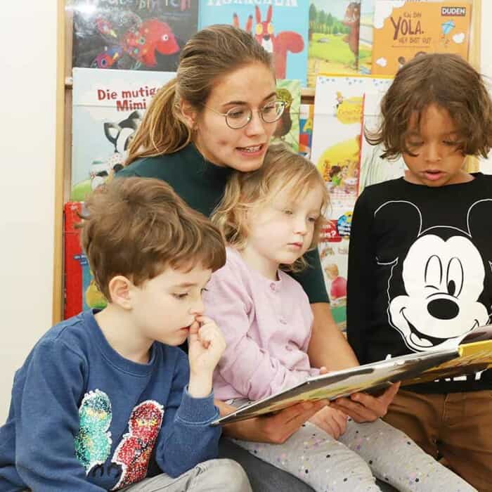 Eine Pädagogin liest ein Buch vor. Ein Mädchen sitzt auf ihrem Schoß, während zwei Jungen daneben stehen.