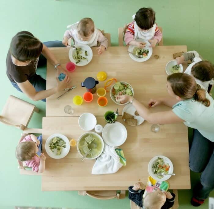 Zwei Erzieher sitzen mit fünf Kinder an einem Tisch. Jedes Kind hat einen Teller mit einem Salat vor sich. Eine Pädagogin beugt sich nach vorne, um sich Salat zu holen.