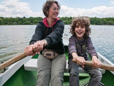 Eine Frau mit Brille und ein Junge mit langen Haaren sitzen in einem Ruderboot auf einem See und rudern lachend.