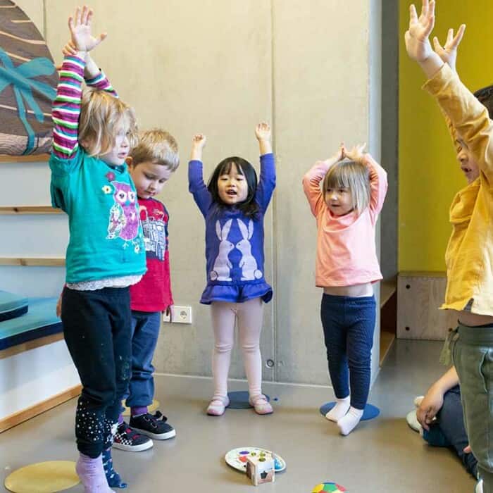 Eine Gruppe Kinder stehen im Kreis, lachen und heben erfreut ihre Arme in die Luft. Eine Erzieherin mit Brille sitzt auf dem Boden.