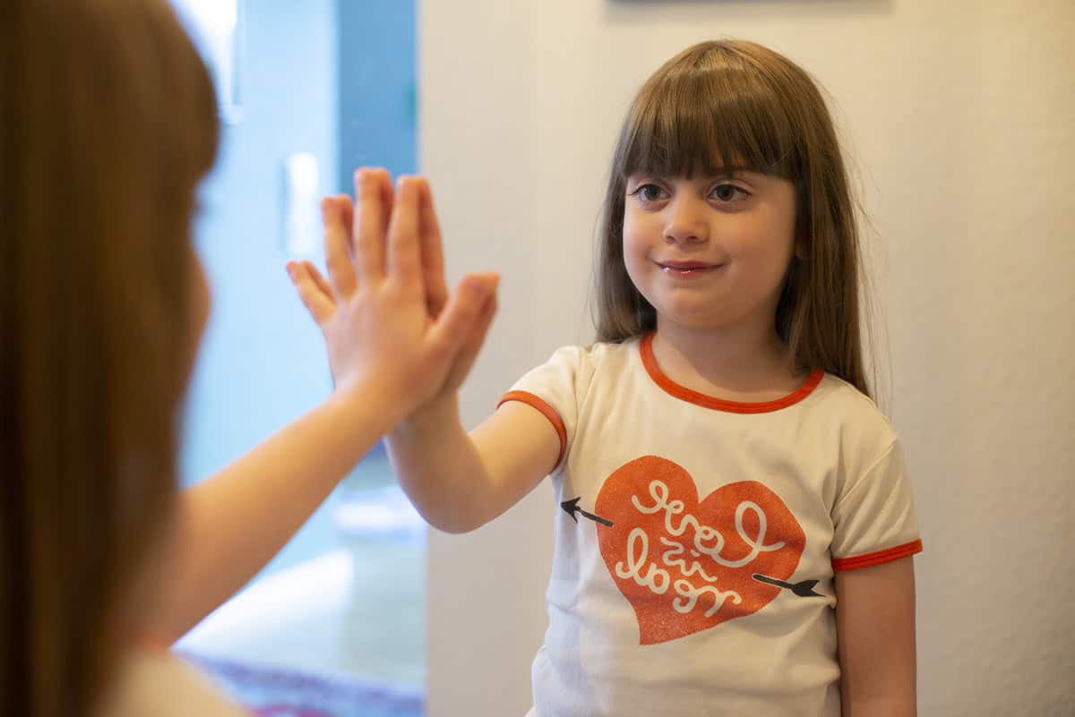 Ein braunhaariges Mädchen steht an einem Spiegel, betrachtet sich und streckt die Hand an den Spiegel nach ihrem Spiegelbild aus.