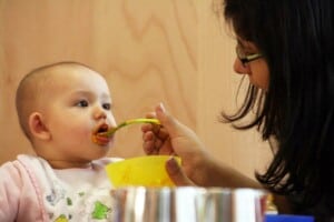 Ein Kleinkind wird von einer FRau mit BRille und dunklen Haaren gefüttert.