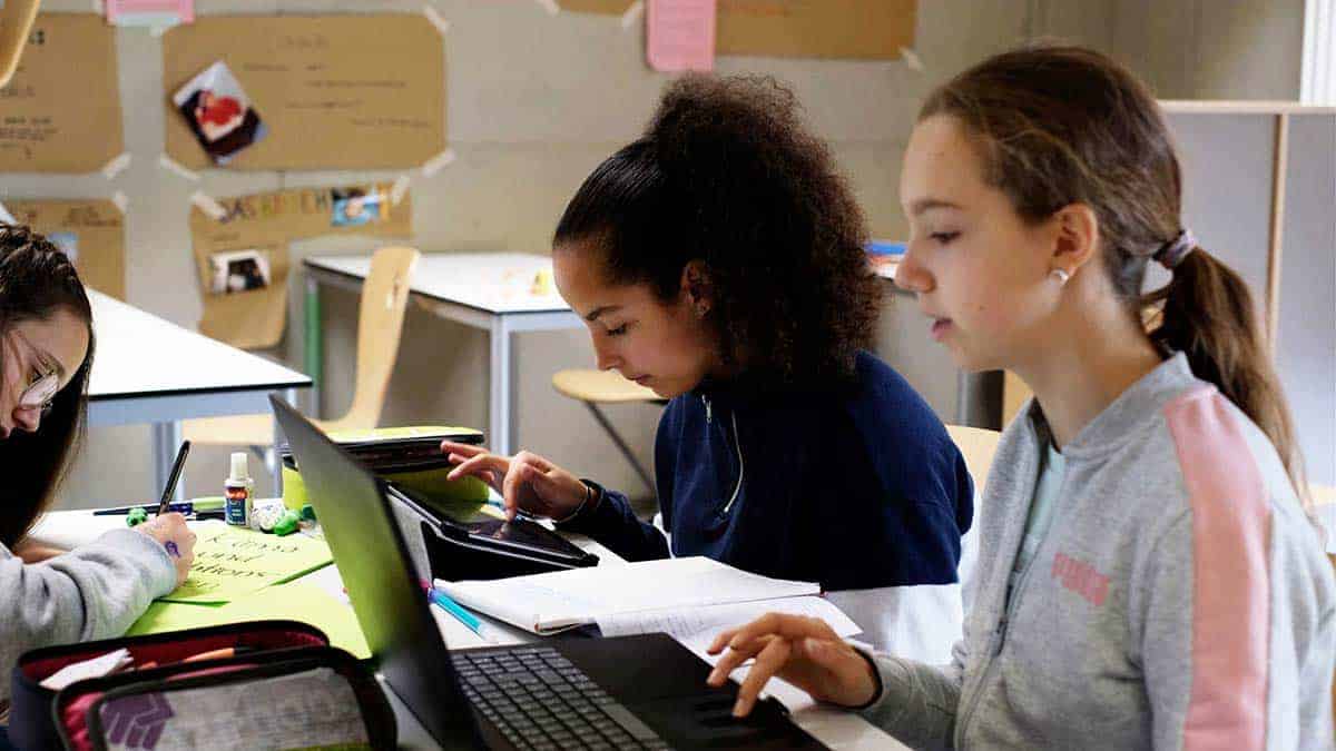 Drei Schülerinnen sitzen zusammen und arbeiten mit Stift und Papier, Tablet und Laptop.