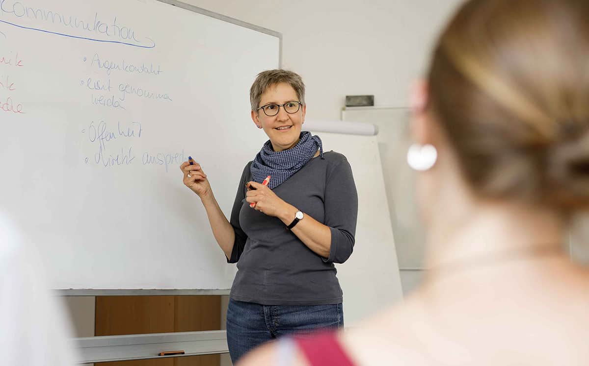 Eine Lehrerin mit Brille steht mit einem Lächeln am Whiteboard / an der Tafel und erklärt etwas.