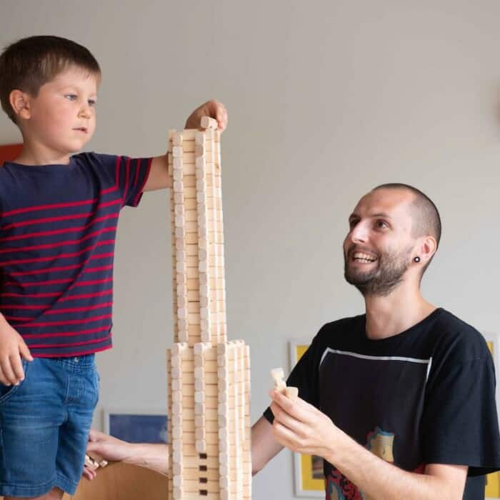 Ein Junge baut einen Turm aus Holzbausteinen und ein lachender Mann sitzt daneben und beobachtet ihn.