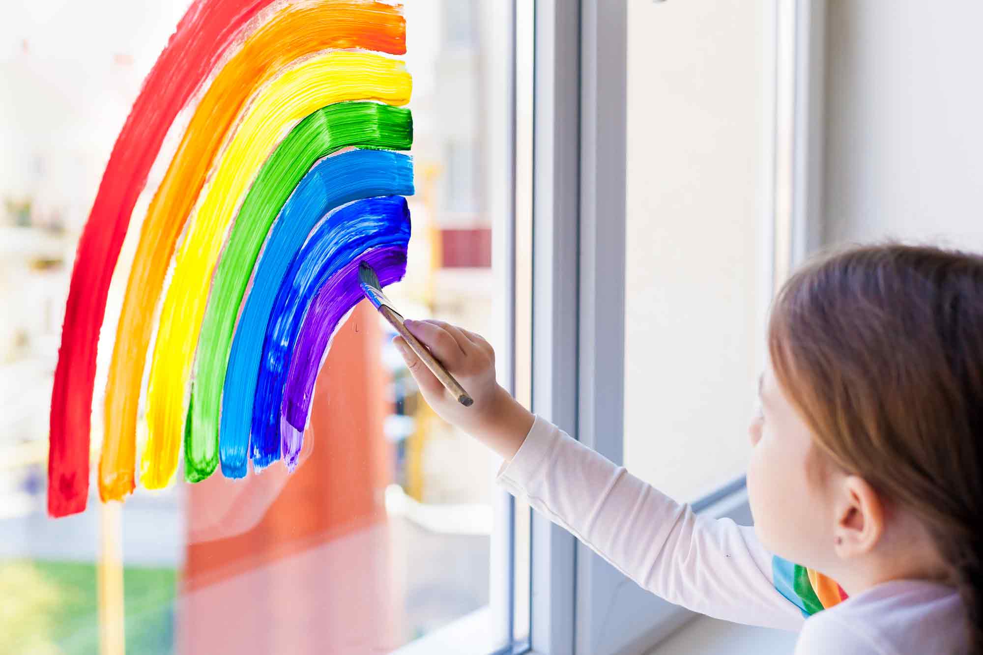 Ein Mädchen malt mit einem Pinsel einen Regenbogen an ein Fenster.