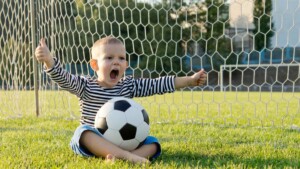 Ein Junge sitzt auf dem Rasen vor einem Tor, hat einen Fußball auf dem Schoß und reißt in Gewinnermanier die Arme auseinander.