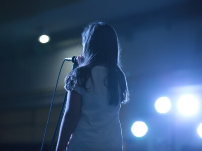 Mädchen spricht oder singt in ein Mikrofon auf der Bühne. Auf sie sind Scheinwerfer gerichtet.
