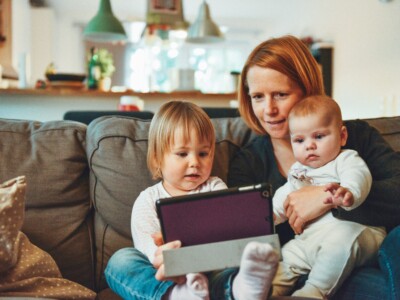 Eine Mutter sitzt mit ihrem Baby und ihrem Kleinkind auf dem Sofa, alle schauen auf ein Tablet.