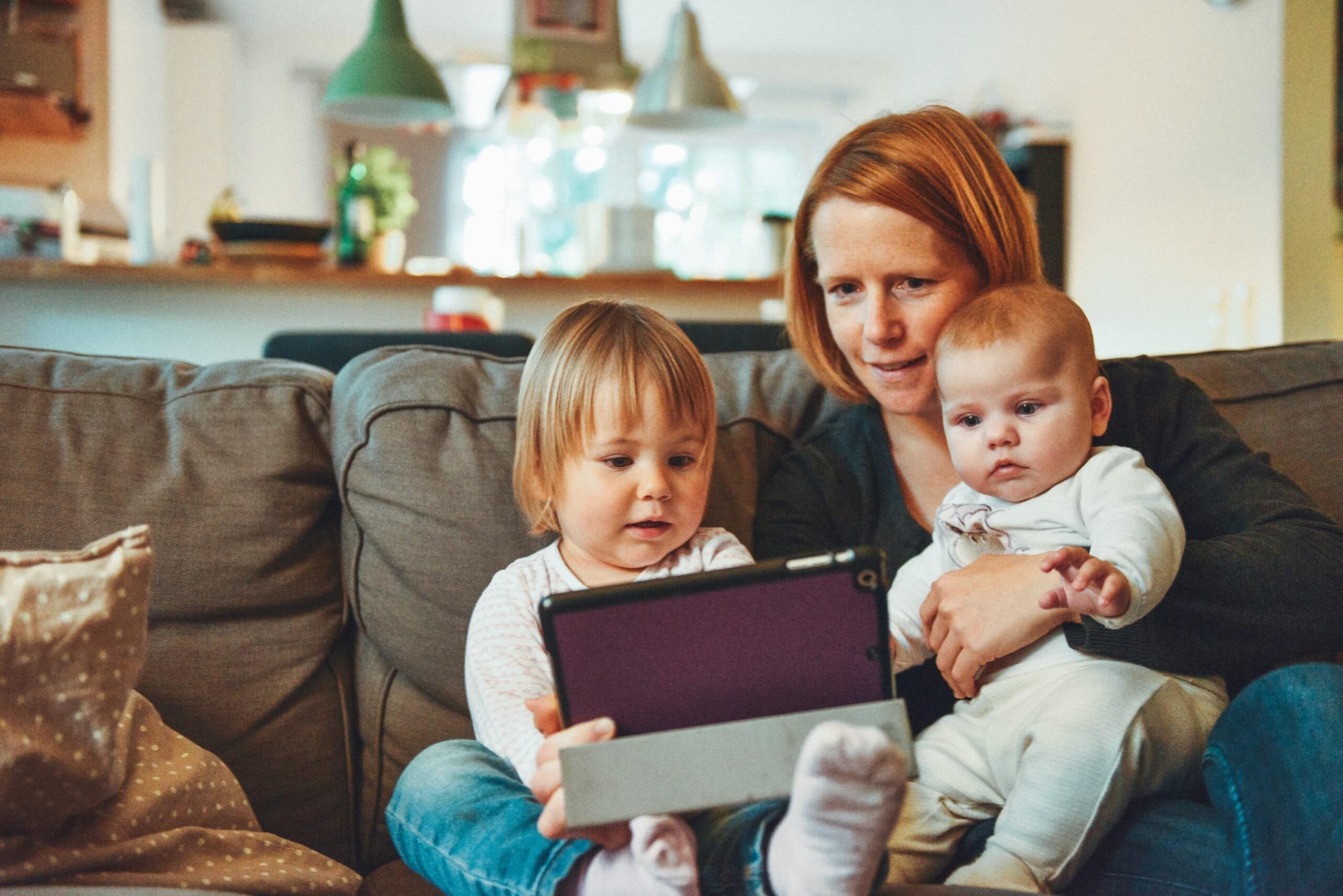 Eine Mutter sitzt mit ihrem Baby und ihrem Kleinkind auf dem Sofa, alle schauen auf ein Tablet.