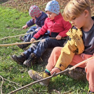 Drei Kinder sitzen auf einer Wiese und spielen mit Stöcken.