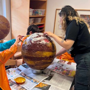 Eine Erzieherin und einige Kinder bemalen einen Pappmaché Planeten.