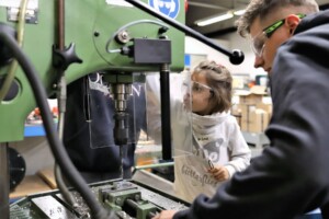 Ein Erwachsener und ein Kind tragen Schutzbrillen. Der Erwachsene zeigt dem Kind eine Bohrmaschine.