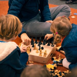 Zwei Kinder und eine Person sitzen auf dem Boden und spielen Schach