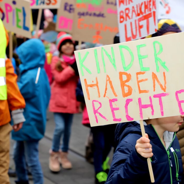 Kinder und Erwachsene auf einer Demonstration für Kinderrechte. Ein Kind zeigt ein Schild mit dem Slogan "Kinderhaben Rechte"