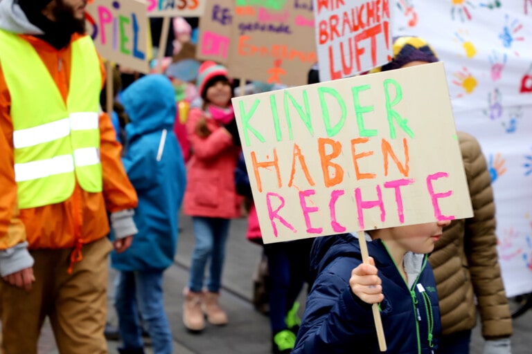 Kinder und Erwachsene auf einer Demonstration für Kinderrechte. Ein Kind zeigt ein Schild mit dem Slogan "Kinder haben Rechte"