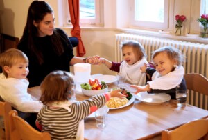 Eine Pädagogin sitzt mit einer Gruppe Kindern am Tisch, das Essen steht auf dem Tisch und alle halten sich an den Händen.