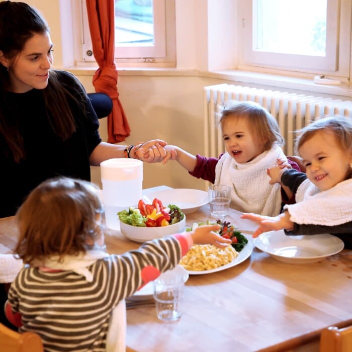 Eine Pädagogin sitzt mit einer Gruppe Kindern am Tisch, das Essen steht auf dem Tisch und alle halten sich an den Händen.